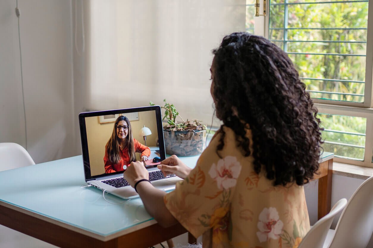 Mulher em home office participando de sessão de apoio psicológico online por videochamada, sentada à mesa com notebook em ambiente residencial iluminado.