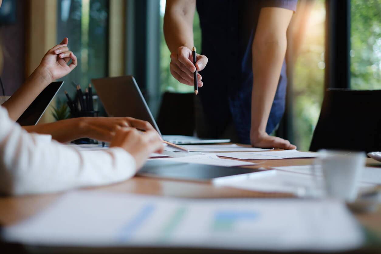 Pessoas em reunião de trabalho organizando tarefas com papéis, canetas e notebooks sobre a mesa, representando planejamento e gestão de tempo em equipe.