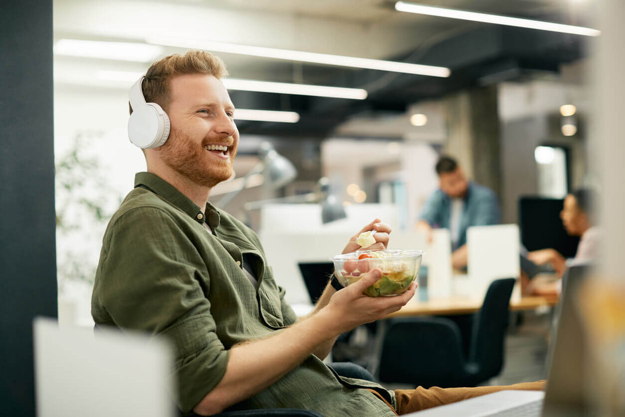 Homem ruivo sentado em ambiente de trabalho, sorrindo enquanto se alimenta com uma salada, simbolizando a importância da alimentação saudável como aliada na prevenção de doenças ocupacionais.