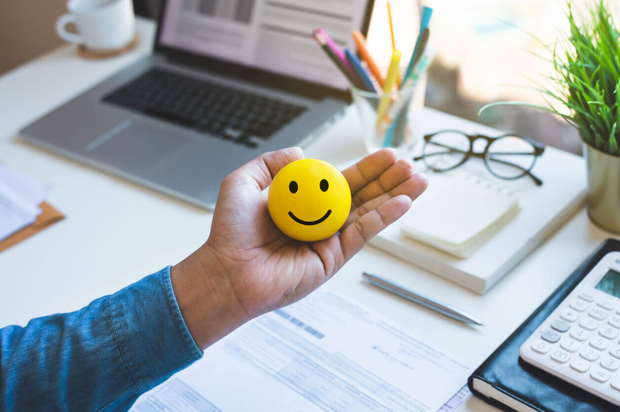Pessoa segurando um emoji amarelo sorridente em ambiente de trabalho, simbolizando bem-estar e saúde mental corporativa como certificado de saúde mental.