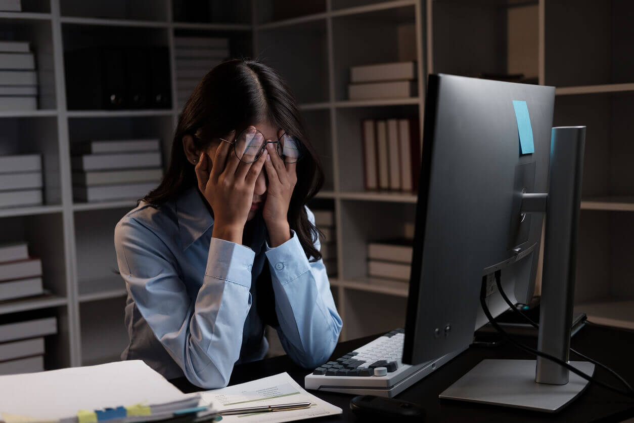 Mulher sentada à mesa de trabalho com as mãos no rosto, demonstrando sinais de estresse ou exaustão em frente ao computador em um escritório.