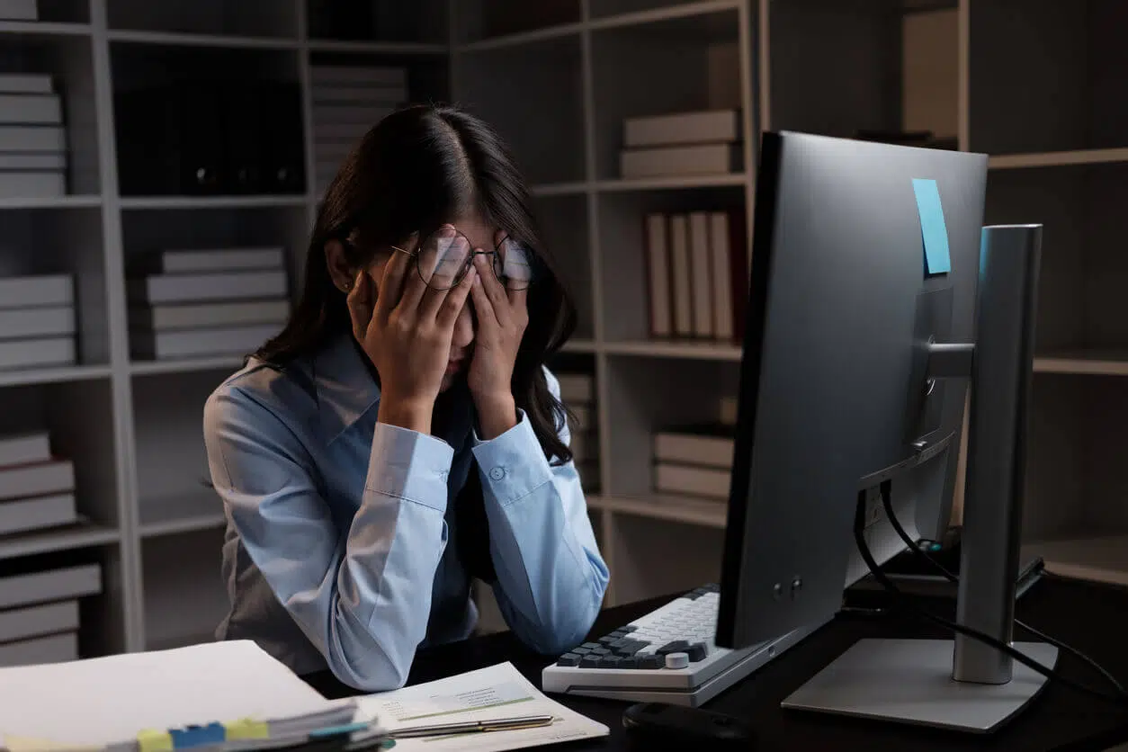 Mulher sentada à mesa de trabalho com as mãos no rosto, demonstrando sinais de estresse ou exaustão em frente ao computador em um escritório.