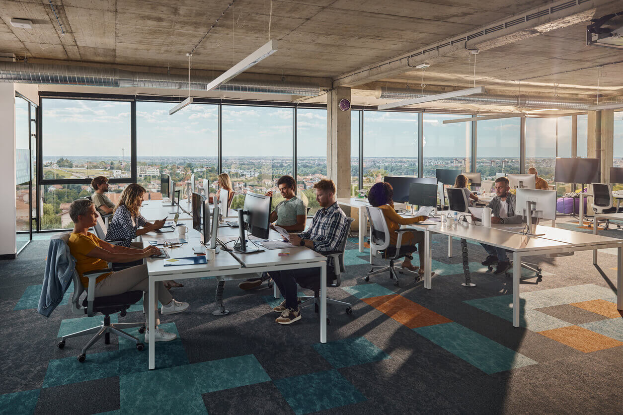 Ambiente corporativo moderno com colaboradores trabalhando em estações de trabalho compartilhadas, representando a importância do cumprimento da nova NR-1 em escritórios e espaços administrativos.