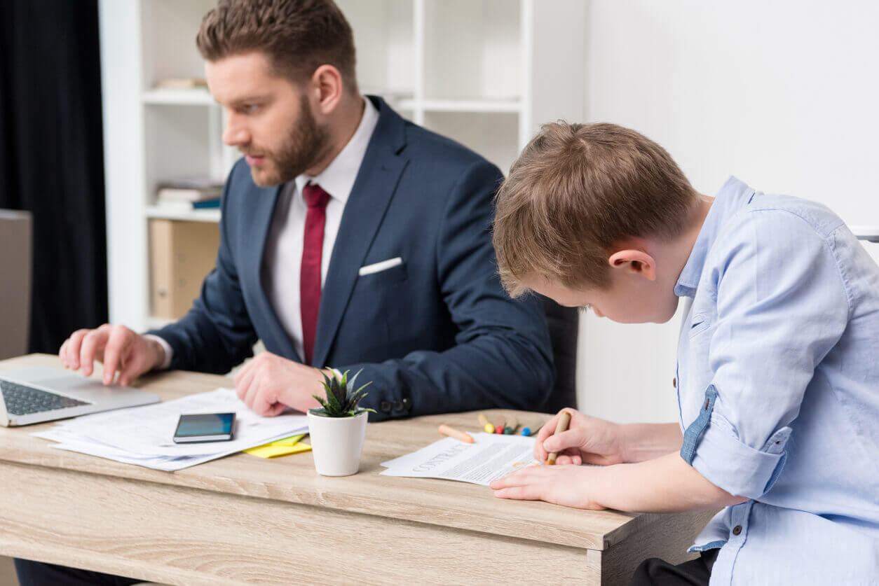 Pai de terno trabalhando em escritório ao lado do filho que desenha sobre a mesa, representando a importância do equilíbrio familiar em ações de endomarketing para o Dia dos Pais.