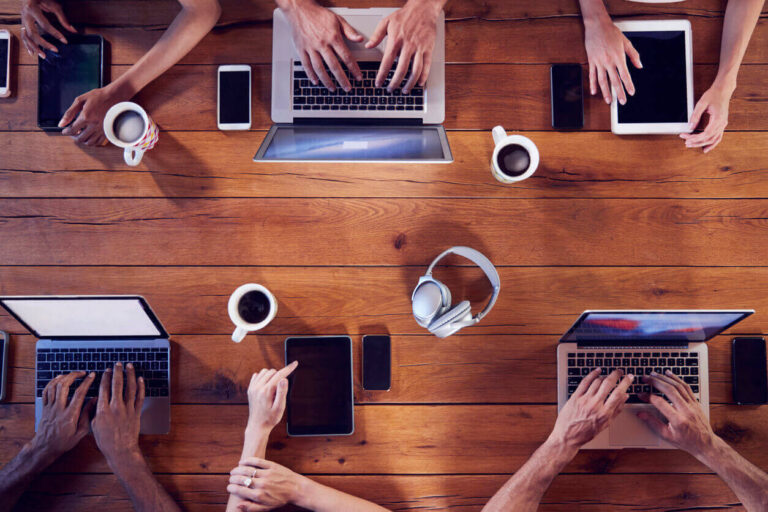 Vista aérea de pessoas trabalhando em laptops e tablets sobre uma mesa de madeira, com smartphones, fones de ouvido e xícaras de café, representando os efeitos da hiperconectividade no ambiente de trabalho