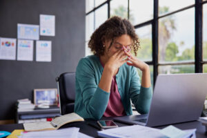 Mulher no escritório com expressão de cansaço e mãos no rosto, representando os sinais da síndrome de Brownout no trabalho
