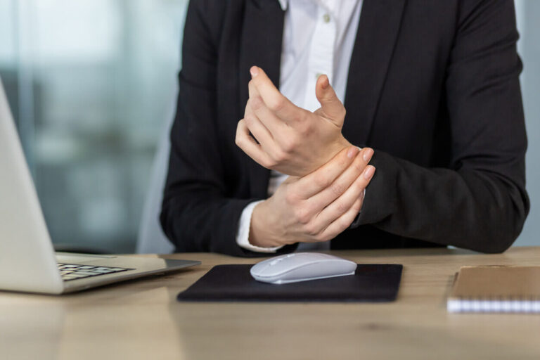 Profissional sentada ao computador segurando o punho com expressão de dor, representando sintomas de LER/DORT e a importância da prevenção no ambiente de trabalho.