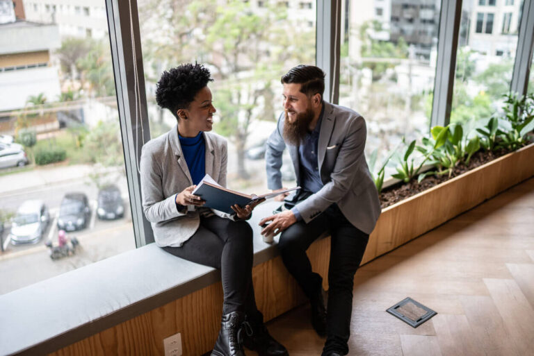 Gestora conversando com colaborador durante um feedback, em ambiente corporativo com vista para a cidade, simbolizando diálogo aberto e fortalecimento do clima organizacional.
