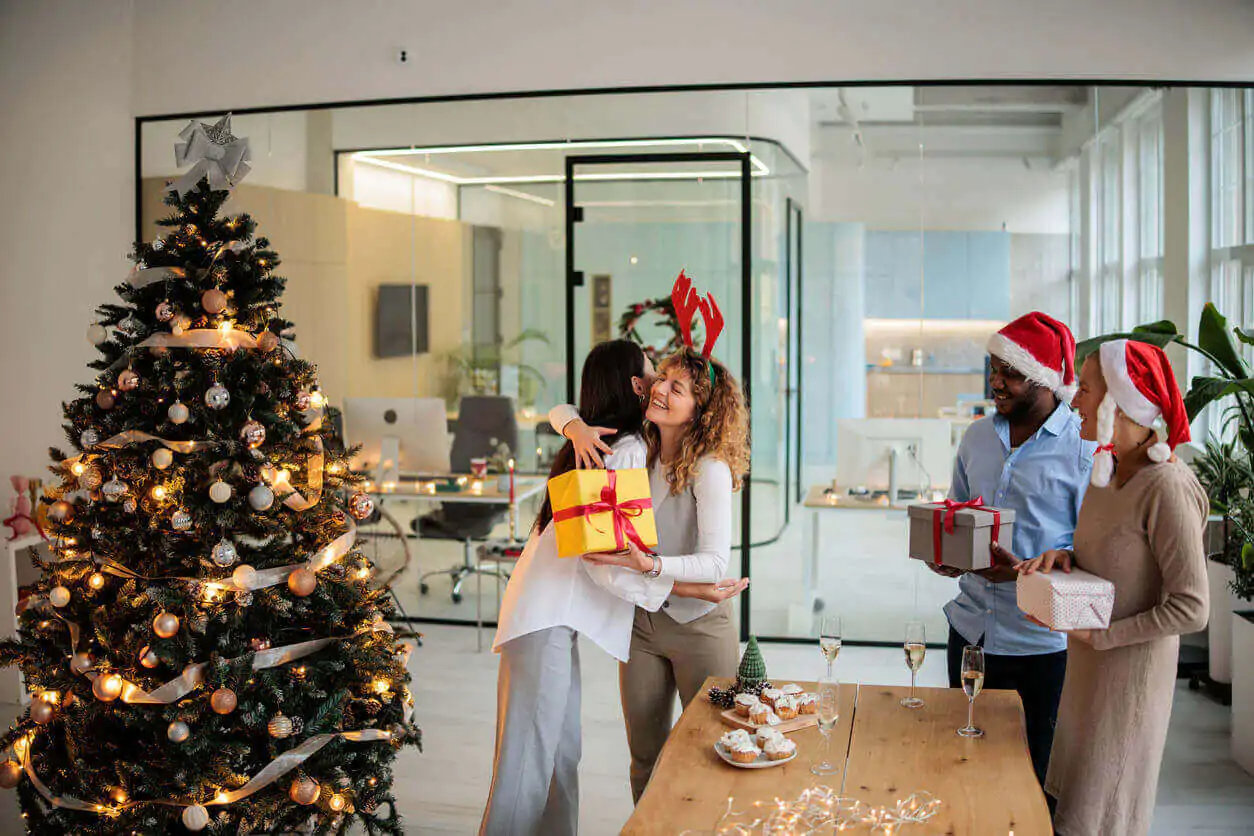 Colaboradores celebrando no escritório, trocando presentes e participando de um amigo secreto típico de confraternização de fim de ano, ao lado da árvore de Natal decorada.