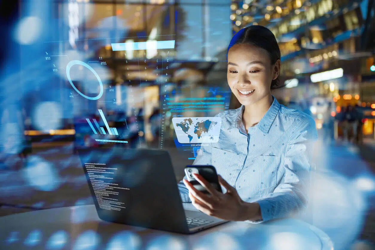Profissional sorridente utilizando notebook e celular em ambiente corporativo, com gráficos digitais refletidos ao redor, representando o uso da tecnologia e da nova NR1 apoiada por inteligência artificial para aprimorar a gestão de segurança no trabalho.