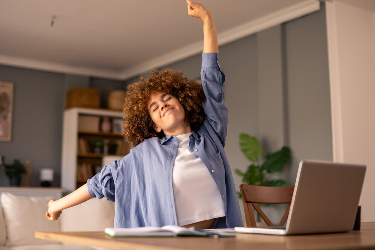 Pessoa sorridente se espreguiçando durante o trabalho remoto, simbolizando bem-estar e saúde mental no home office, com notebook e caderno sobre a mesa em um ambiente aconchegante.