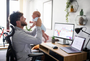 Pai trabalhando em home office enquanto segura o bebê no colo, simbolizando o desafio e a busca por equilíbrio entre vida pessoal e trabalho.