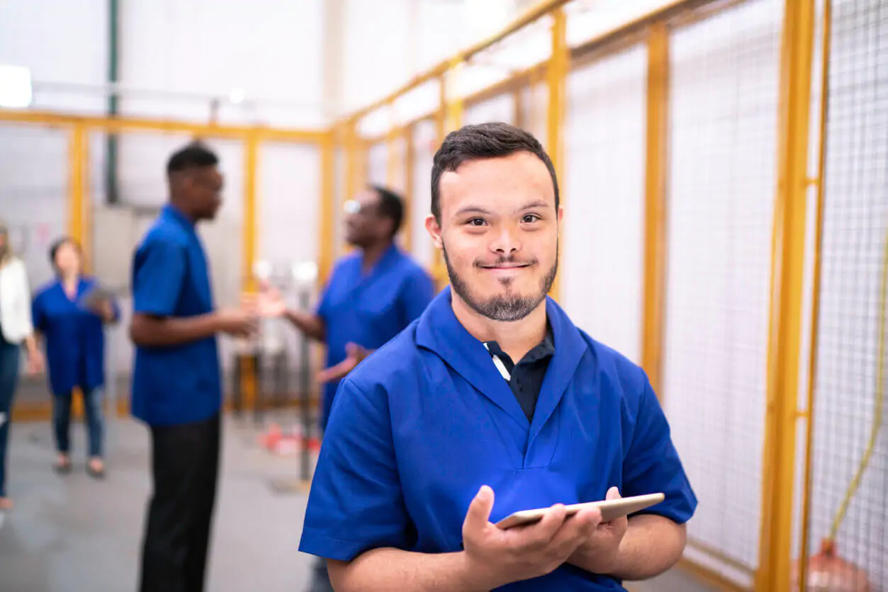 Jovem trabalhador com Síndrome de Down sorrindo e segurando um tablet em um ambiente de fábrica ou oficina, com outros colegas ao fundo, simbolizando a importância da acessibilidade e inclusão no mercado de trabalho.